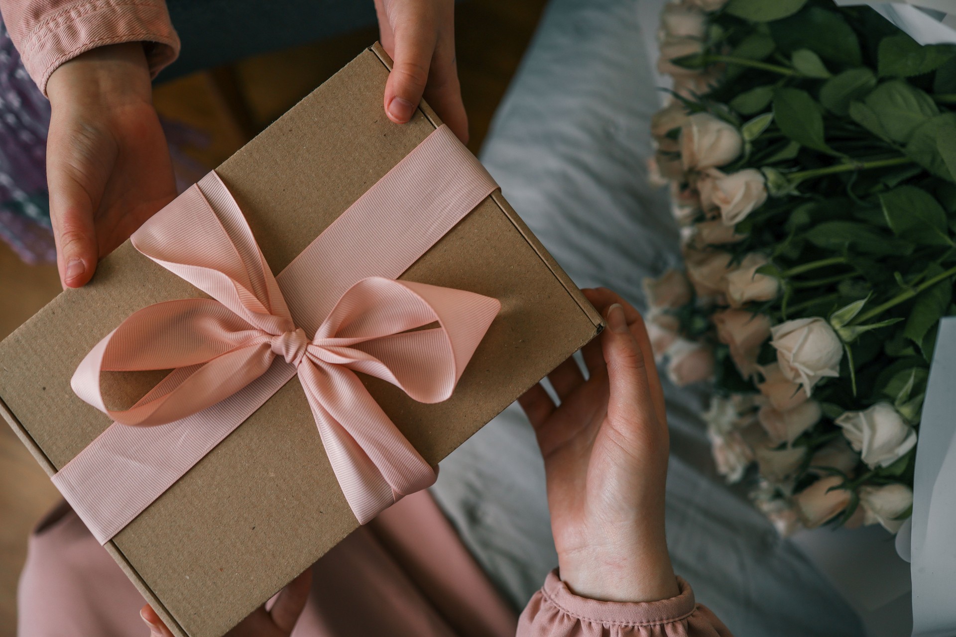 Hands of a child giving a wrapped gift with a satin ribbon to a woman holding a bouquet of roses, captured in warm indoor light